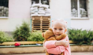 Flicka i Ukraina står framför ett skadat hus och kramar en nalle.