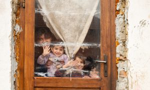 En mamma och tre barn står och vinkar genom ett fönster i ett hus i dåligt skick.