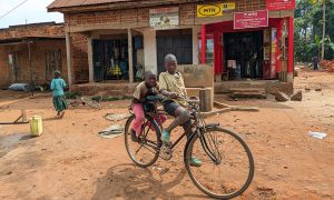 Enkelt hus med två små kiosker/butiker. I förgrunden två barn på en cykel.